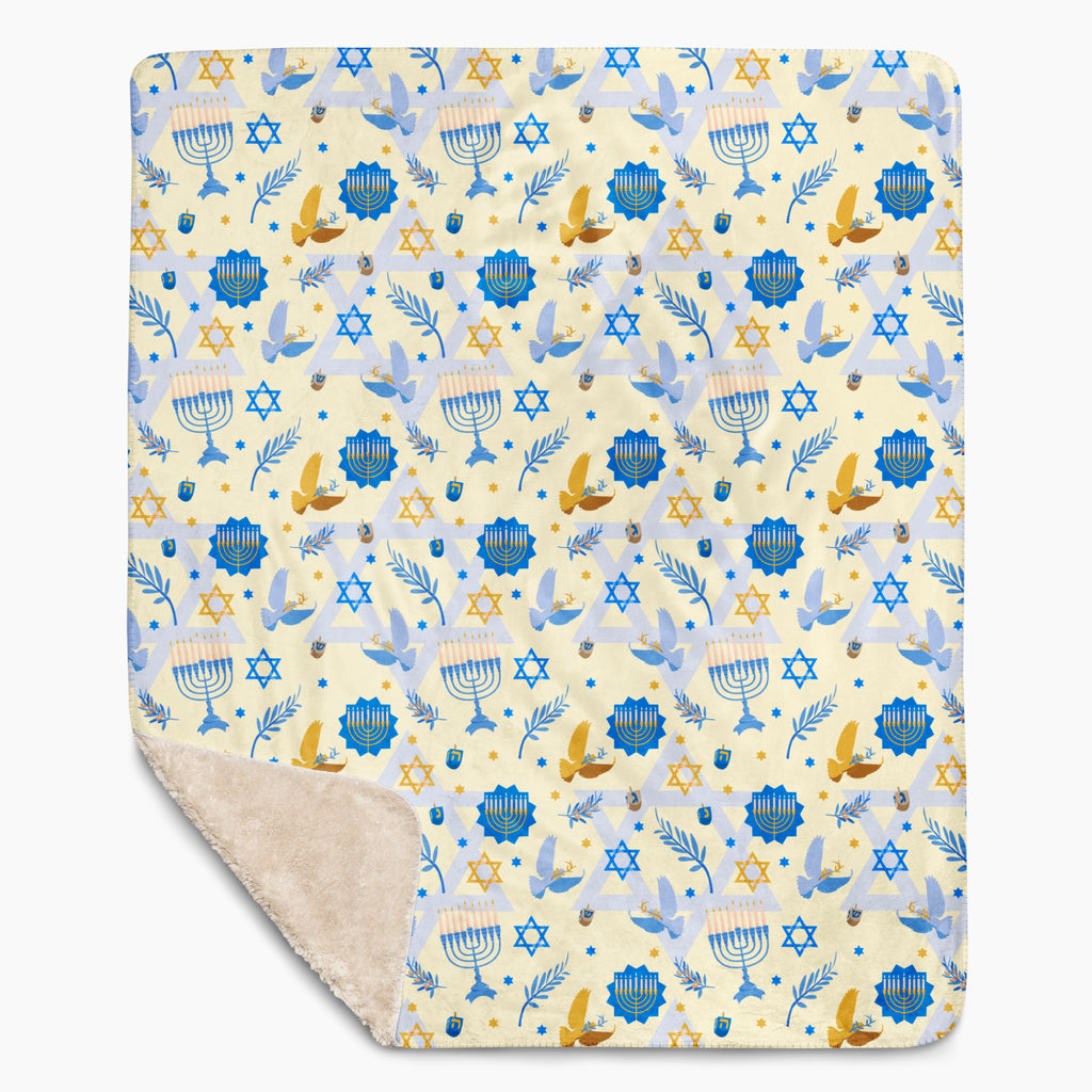 A rectangular blanket with a cream-colored sherpa lining and a printed front featuring a festive pattern of blue and gold menorahs, Star of David, dreidels, doves, and olive branches on a pale yellow background.