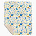 A rectangular blanket with a cream-colored sherpa lining and a printed front featuring a festive pattern of blue and gold menorahs, Star of David, dreidels, doves, and olive branches on a pale yellow background.