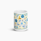 A white ceramic mug with a festive Hanukkah-themed print featuring blue Star of David, Menorahs, doves, and dreidels on a pale yellow background, set against a clean light grey studio backdrop.