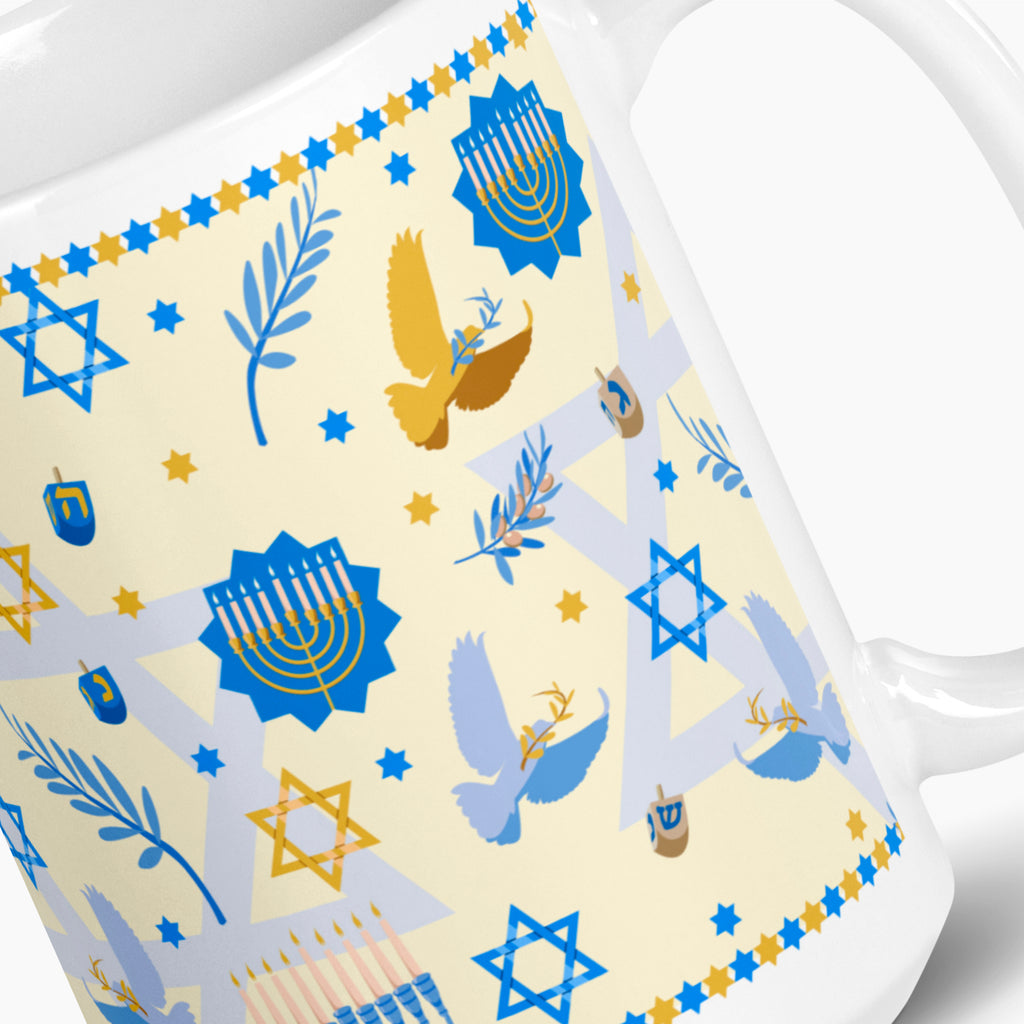 A close-up of a white ceramic mug featuring a festive Hanukkah pattern with blue and gold illustrations of menorahs, dreidels, stars, doves, and olive branches on a cream/pale yellow background.