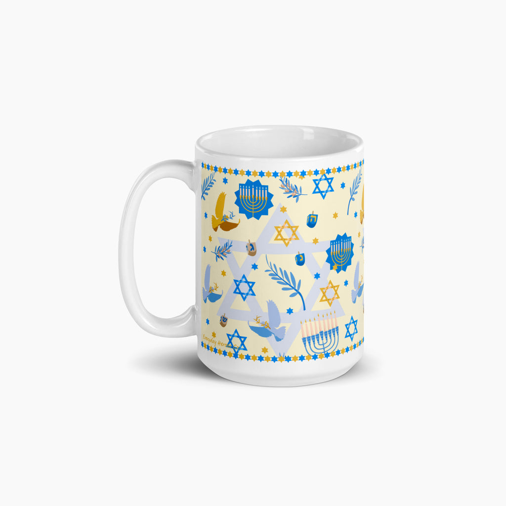 A white ceramic mug with a festive Hanukkah-themed print featuring blue Star of David, Menorahs, doves, and dreidels on a pale yellow background, set against a clean light grey studio backdrop.