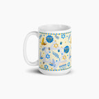 A white ceramic mug with a festive Hanukkah-themed print featuring blue Star of David, Menorahs, doves, and dreidels on a pale yellow background, set against a clean light grey studio backdrop.