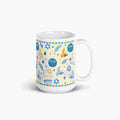 A white ceramic mug with a festive Hanukkah-themed print featuring blue and gold illustrations of menorahs, dreidels, stars, and golden and blue doves on a pale yellow background.