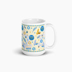 A white ceramic mug with a festive Hanukkah-themed print featuring blue and gold illustrations of menorahs, dreidels, stars, and golden and blue doves on a pale yellow background.