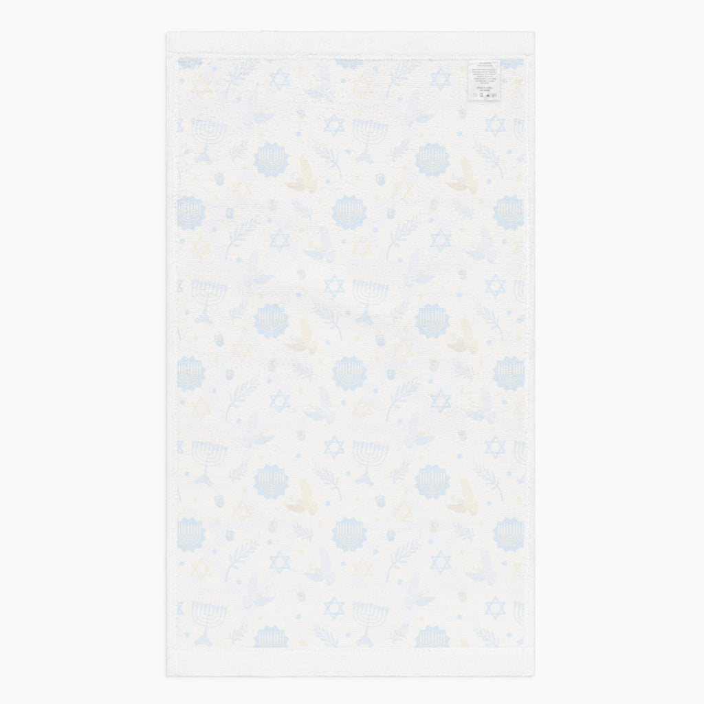 The white backside of a rectangular microfiber fabric hand towel with a repeating festive pattern featuring blue menorahs, blue Star of David motifs, blue circular medallions, gold stars, and small gold and blue doves shown flat against a light grey background