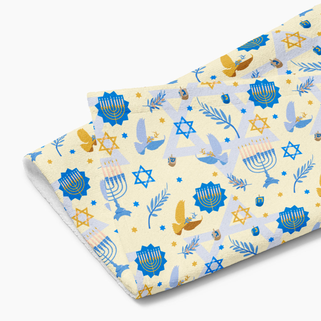 A folded cream-colored fabric hand towel with a festive Hanukkah pattern featuring blue menorahs, gold stars, doves, and olive branches, displayed on a clean light grey background.