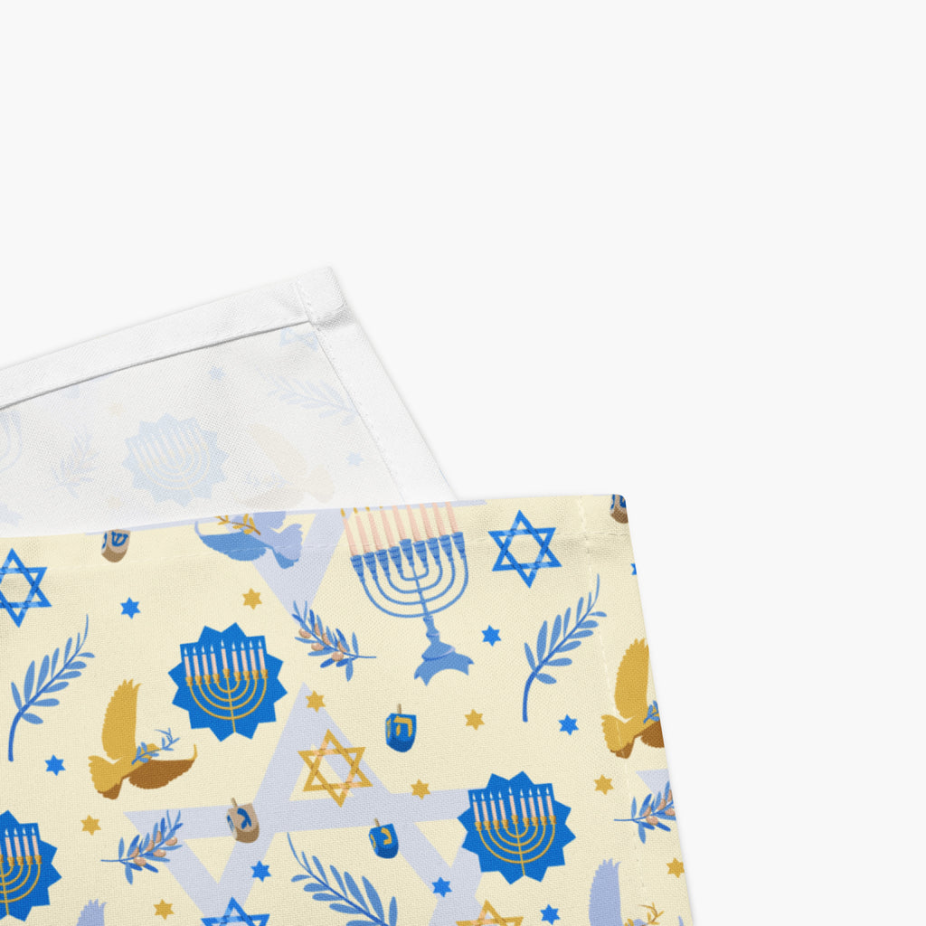 A close-up of folded cloth napkin with a Hanukkah pattern, featuring a cream background with blue and gold illustrations of a menorah, Star of David, dreidels, olive branches, and doves, with the white fabric underside visible.