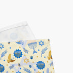 A close-up of folded cloth napkin with a Hanukkah pattern, featuring a cream background with blue and gold illustrations of a menorah, Star of David, dreidels, olive branches, and doves, with the white fabric underside visible.
