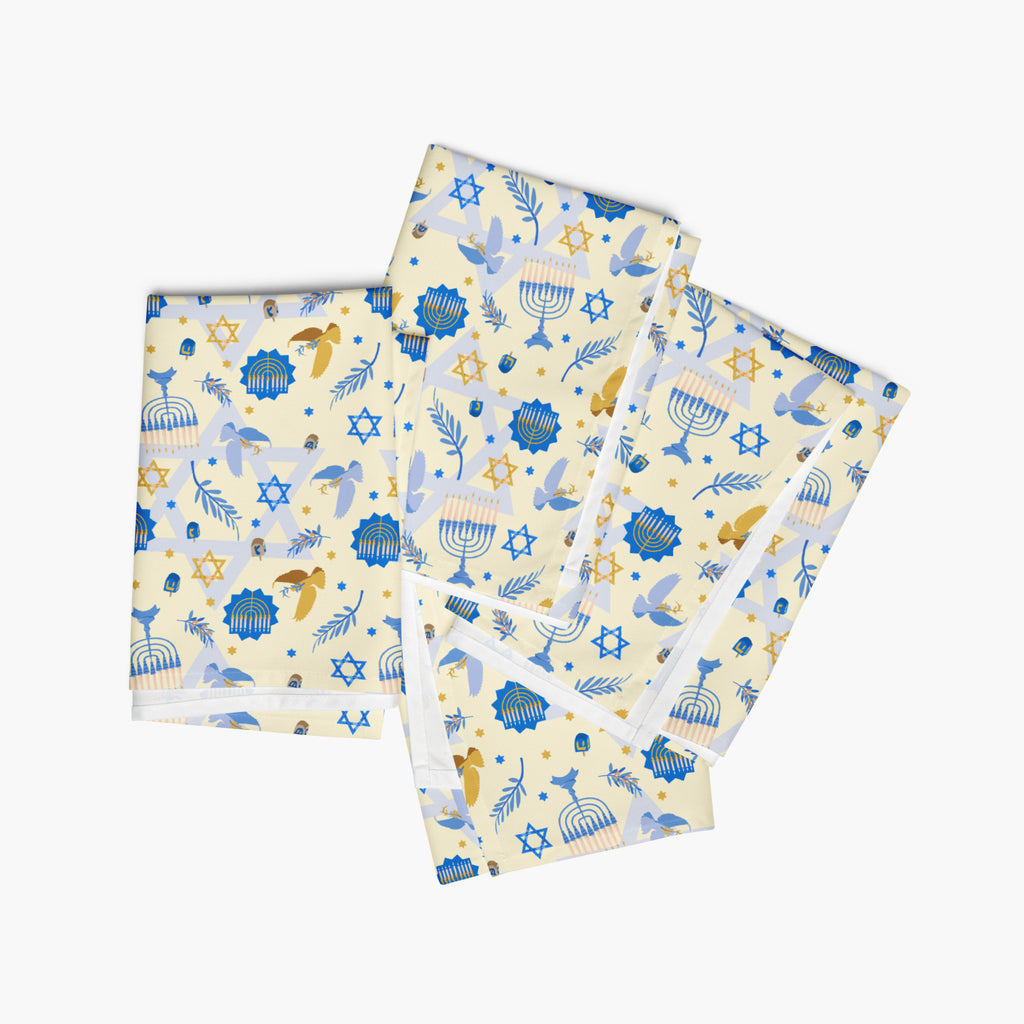 Four folded square cloth napkins with a festive Hanukkah-themed pattern featuring blue Star of David, dreidels, doves, olive branches, and menorahs on a pale yellow background, arranged in a stack on a light gray background.