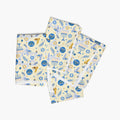 Four folded square cloth napkins with a festive Hanukkah-themed pattern featuring blue Star of David, dreidels, doves, olive branches, and menorahs on a pale yellow background, arranged in a stack on a light gray background.