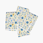 Four folded square cloth napkins with a festive Hanukkah-themed pattern featuring blue Star of David, dreidels, doves, olive branches, and menorahs on a pale yellow background, arranged in a stack on a light gray background.