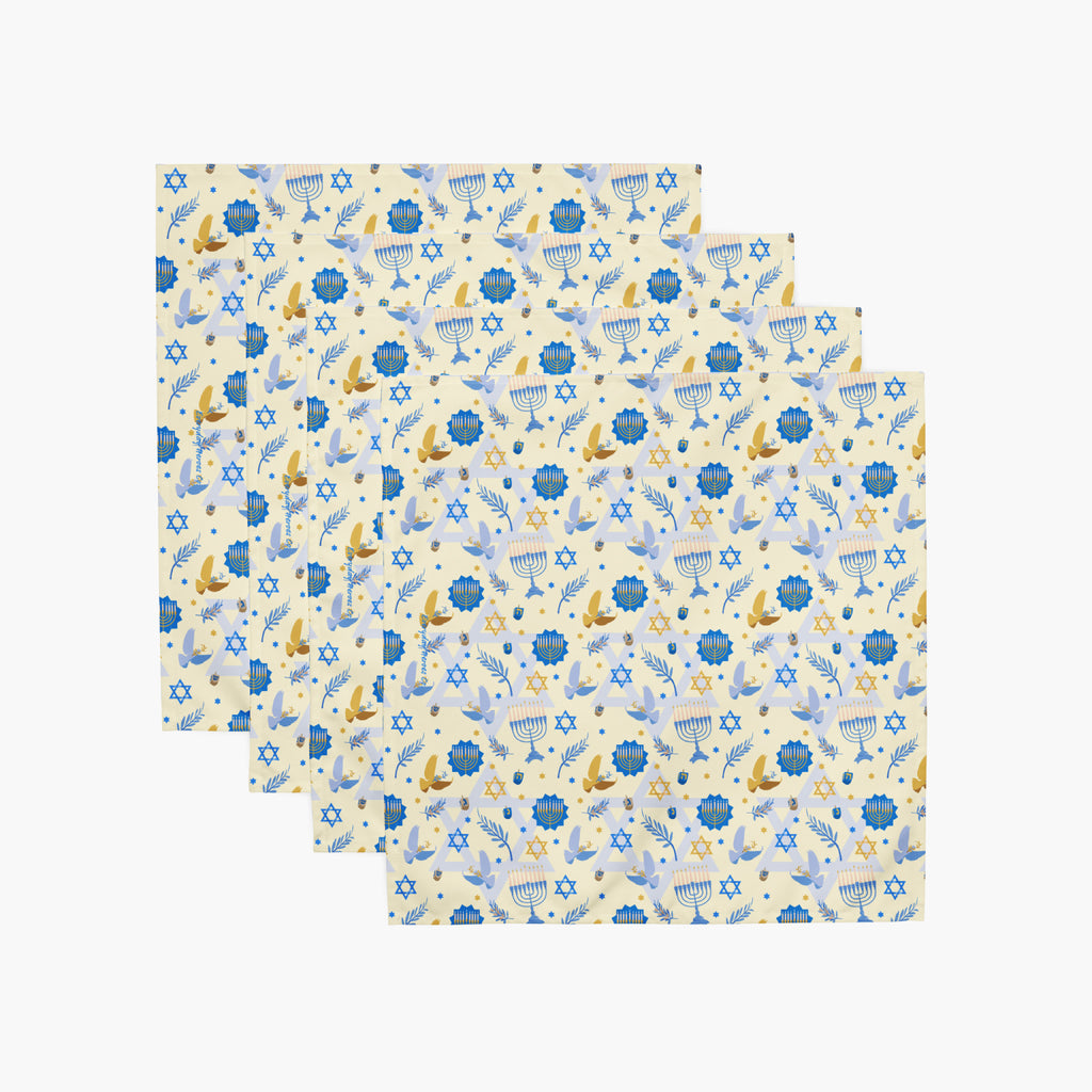 Four square cloth napkins with a festive Hanukkah-themed pattern featuring blue Star of David, dreidels, doves, olive branches, and menorahs on a pale yellow background, arranged in a staggered stack on a light gray background.