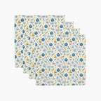 Four square cloth napkins with a festive Hanukkah-themed pattern featuring blue Star of David, dreidels, doves, olive branches, and menorahs on a pale yellow background, arranged in a staggered stack on a light gray background.