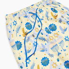 Close-up of a pair of pajama pants with a light cream background and a festive pattern featuring blue menorahs, Star of David, doves, and gold stars. The pants have an elastic waistband and a blue drawstring.