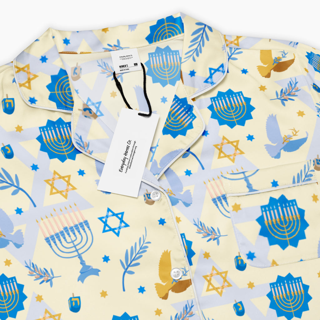A close-up of a pajama top with a festive holiday print featuring blue menorahs, gold Star of David motifs, doves, and blue stars on a pale cream background. The pajama has a white piping border and a white hangtag from 'Everyday Heroes Co'.