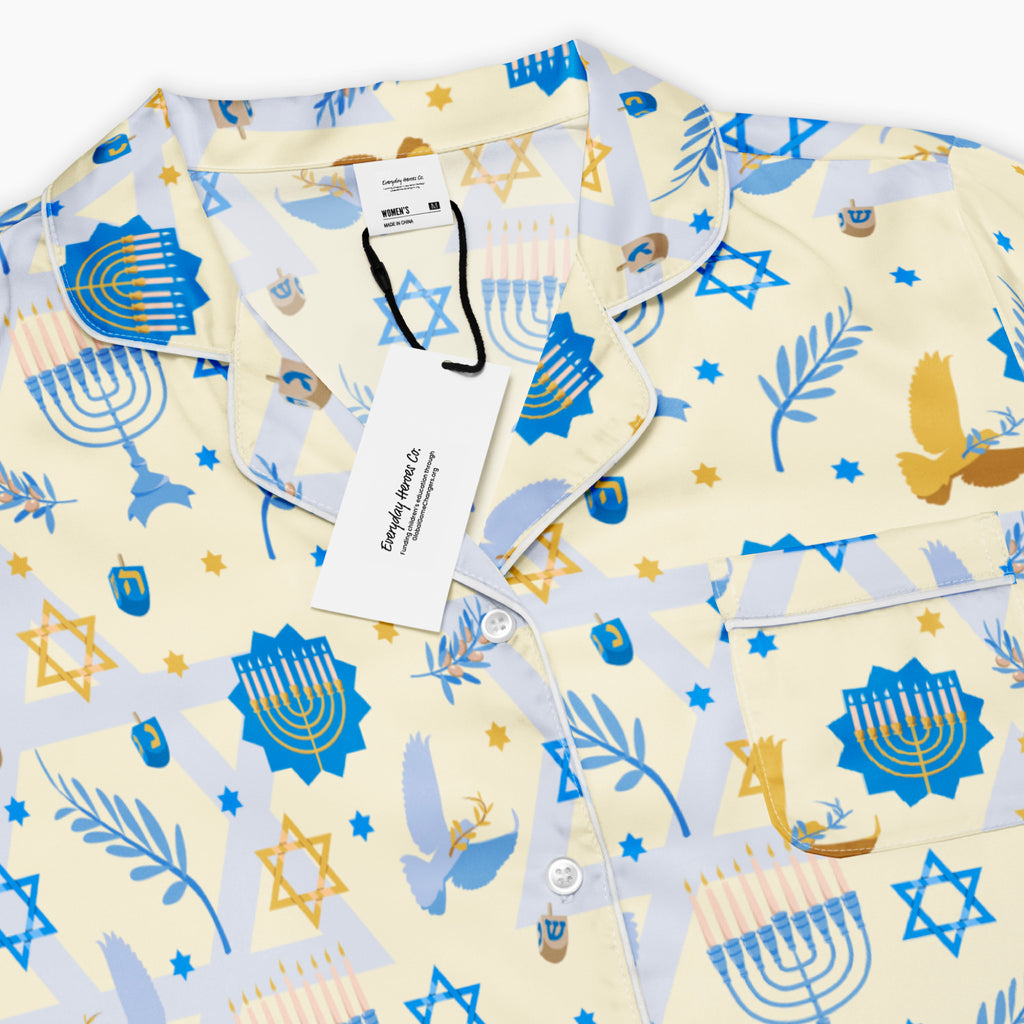A close-up of a short-sleeve button-up pajama shirt with a festive Hanukkah-themed print featuring blue menorahs, Star of David emblems, lulav and olive branches and gold and blue doves on a pale yellow background.
