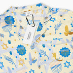 A close-up of a short-sleeve button-up pajama shirt with a festive Hanukkah-themed print featuring blue menorahs, Star of David emblems, lulav and olive branches and gold and blue doves on a pale yellow background.