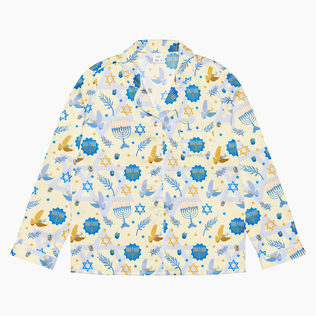 A long-sleeve button-up pajama shirt with a festive Hanukkah-themed print featuring blue menorahs, Star of David emblems, lulav and olive branches and gold and blue doves on a pale yellow background.