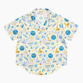A short-sleeve button-up pajama shirt with a festive Hanukkah-themed print featuring blue menorahs, Star of David emblems, lulav and olive branches and gold and blue doves on a pale yellow background.
