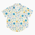 A short-sleeve button-up pajama shirt with a festive Hanukkah-themed print featuring blue menorahs, Star of David emblems, lulav and olive branches and gold and blue doves on a pale yellow background.