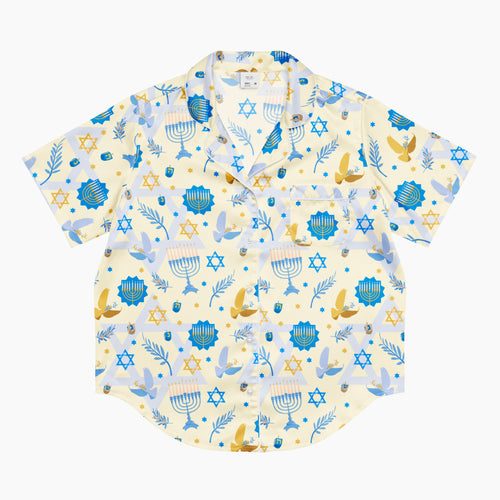 A short-sleeve button-up pajama shirt with a festive Hanukkah-themed print featuring blue menorahs, Star of David emblems, lulav and olive branches and gold and blue doves on a pale yellow background.