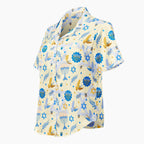 A short-sleeve button-up pajama shirt with a festive Hanukkah-themed print featuring blue menorahs, Star of David emblems, lulav and olive branches and gold and blue doves on a pale yellow background shown from the side against a light grey studio backdrop.
