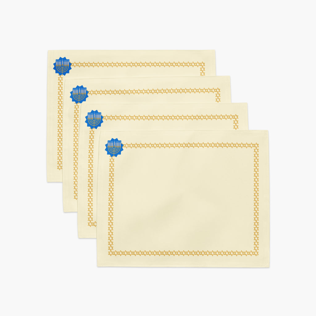 A stack of 4 staggered rectangular cream-colored fabric placemat with a decorative border. The border features a repeating gold Star of David pattern around the edges, and a blue medallion with a gold menorah design in the top-left corner.