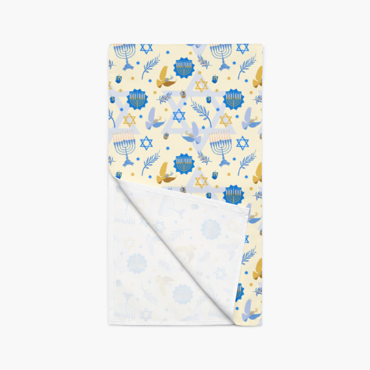 A table runner with a festive Hanukkah-themed print featuring blue menorahs, Star of David emblems, lulav and olive branches and gold and blue doves on a pale yellow background with bottom the corner folded up to show the plain white fabric underside.
