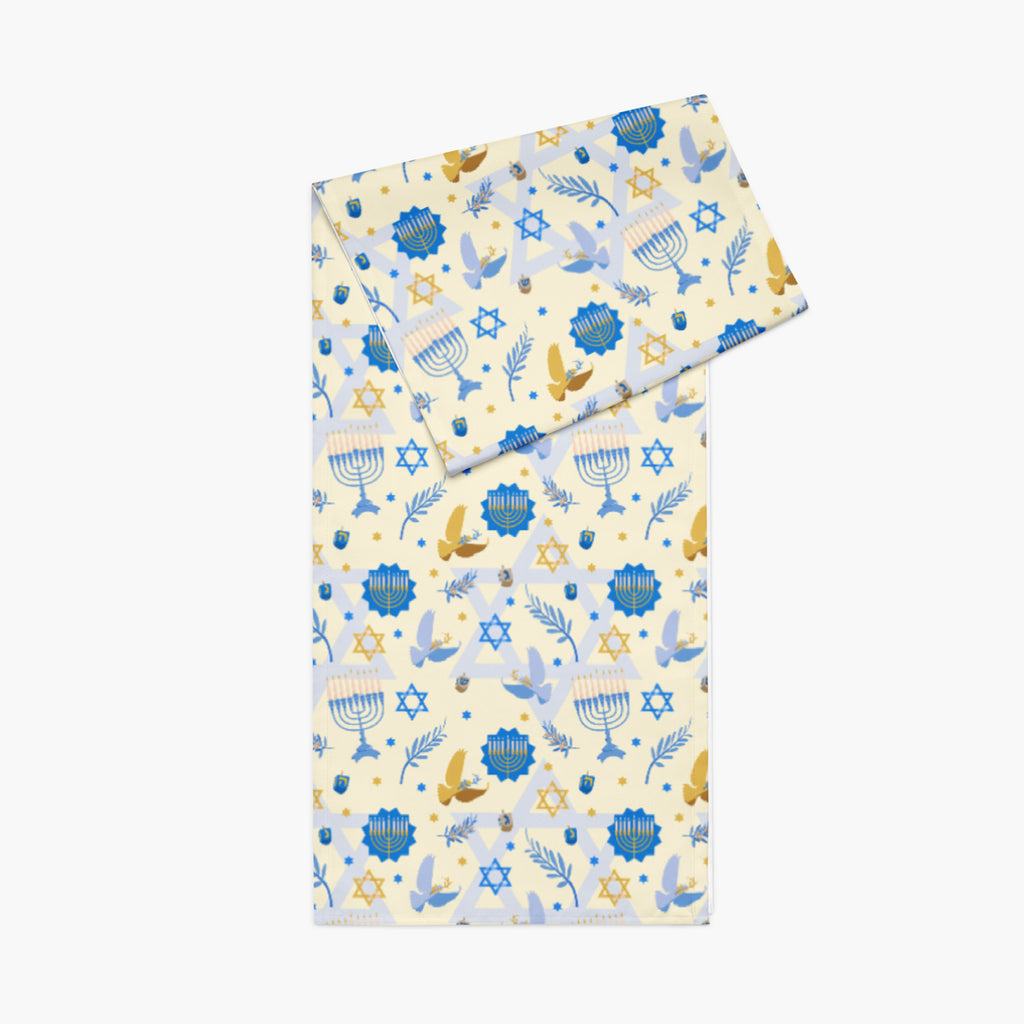 A folded table runner with a festive Hanukkah-themed print featuring blue menorahs, Star of David emblems, lulav and olive branches and gold and blue doves on a pale yellow background.
