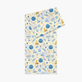 A folded table runner with a festive Hanukkah-themed print featuring blue menorahs, Star of David emblems, lulav and olive branches and gold and blue doves on a pale yellow background.