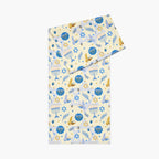A folded table runner with a festive Hanukkah-themed print featuring blue menorahs, Star of David emblems, lulav and olive branches and gold and blue doves on a pale yellow background.