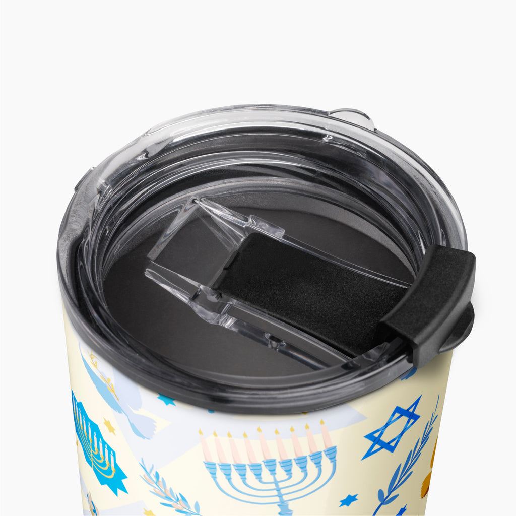 Close-up of a tumbler with a clear lid and black flip-cover component, decorated with blue Hanukkah illustrations including a menorah, dreidel, stars, and olive branches on a cream background.