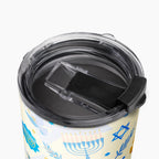 Close-up of a tumbler with a clear lid and black flip-cover component, decorated with blue Hanukkah illustrations including a menorah, dreidel, stars, and olive branches on a cream background.