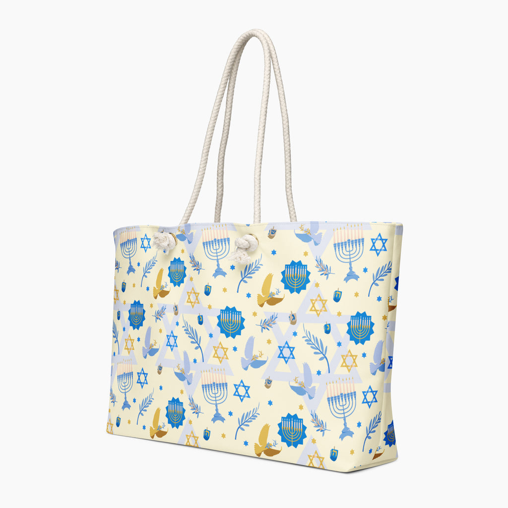 A tote bag with a festive Hanukkah pattern featuring blue menorahs, Star of David, dreidels, and gold and blue doves on a cream background, complete with two thick white rope handles.