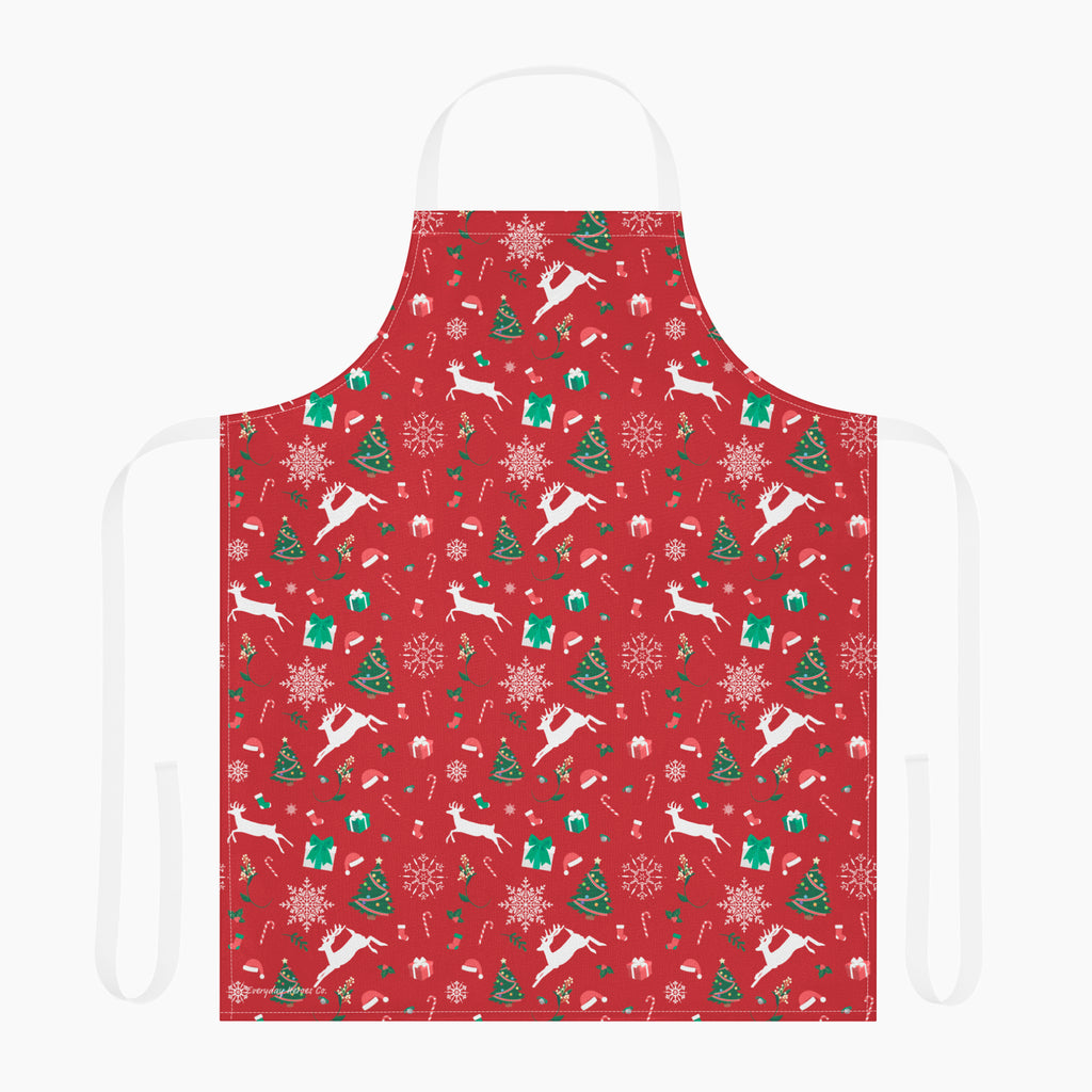 A red apron with a festive Christmas pattern featuring white reindeer, green Christmas trees, snowflakes, gift boxes and candy canes, shown against a plain light grey studio background.