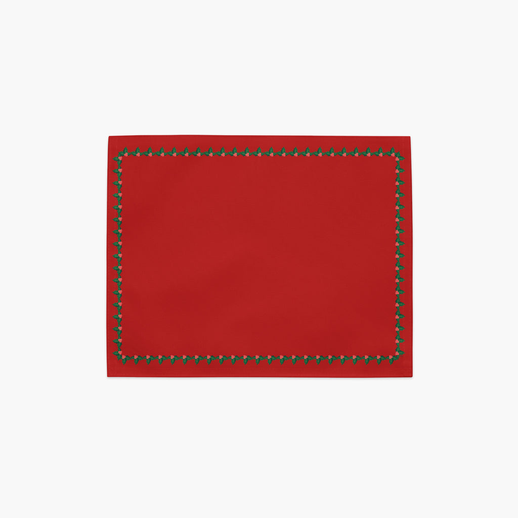 A rectangular red placemat with a decorative border of small green holly leaves and berries shown flat against a light gray background.