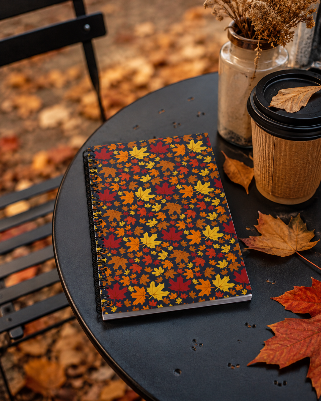 A spiral notebook with a dark background and a dense pattern of colorful autumn maple leaves in red, orange, yellow, and brown, styled on a black outdoor table next to a coffee cup and a glass jar of dried flowers.