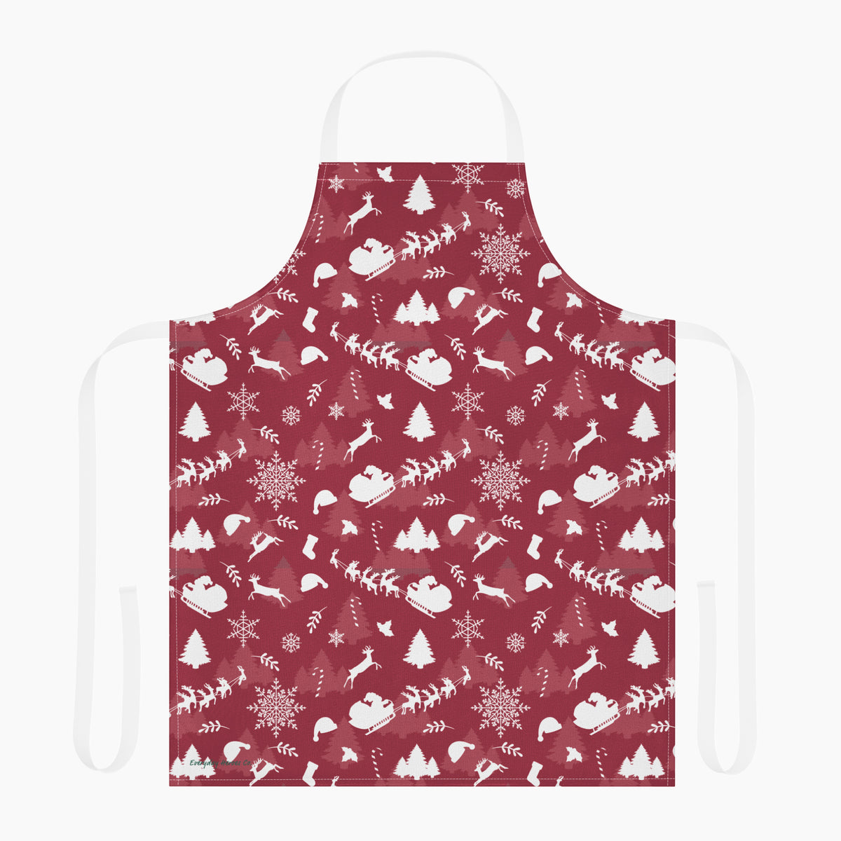 A deep red apron with a white winter holiday pattern featuring snowflakes, Christmas trees, reindeer, sleighs, and mountains, shown against a plain light grey background.