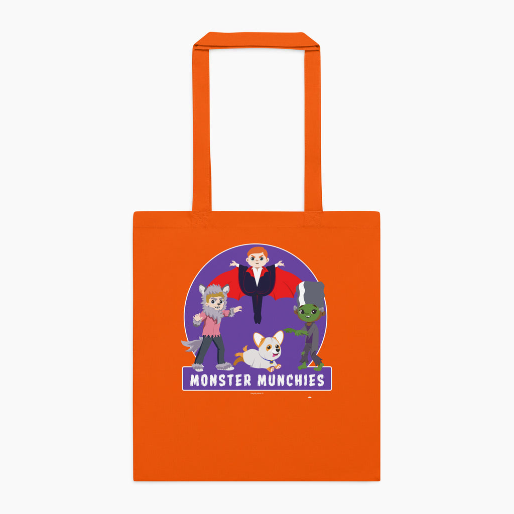 An orange canvas tote bag with orange handles, featuring a colorful graphic print of cartoon monster characters over a purple circle with the text 'MONSTER MUNCHIES' on the front, set against a plain light grey background.