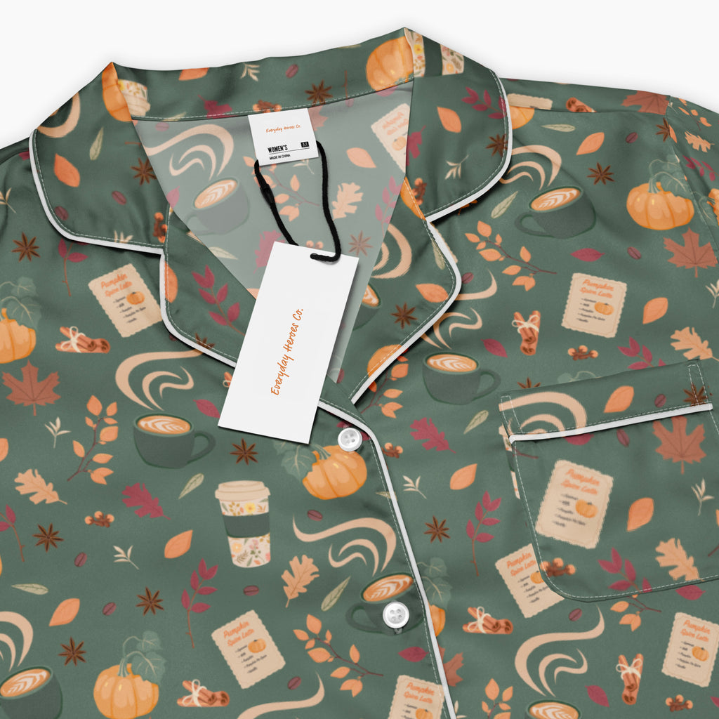 A close-up of a pajama top with a sage green base and an all-over pumpkin spice-themed print featuring coffee cups, pumpkins, leaves, and fall spices. The pajama has white piping along the collar, placket, and pocket, and is shown with a white brand tag and paper labels attached.