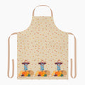 A beige apron with a light tan neck loop and side ties, decorated with a repeating pattern of scattered autumn leaves and three scarecrows standing next to pumpkins at the bottom.
