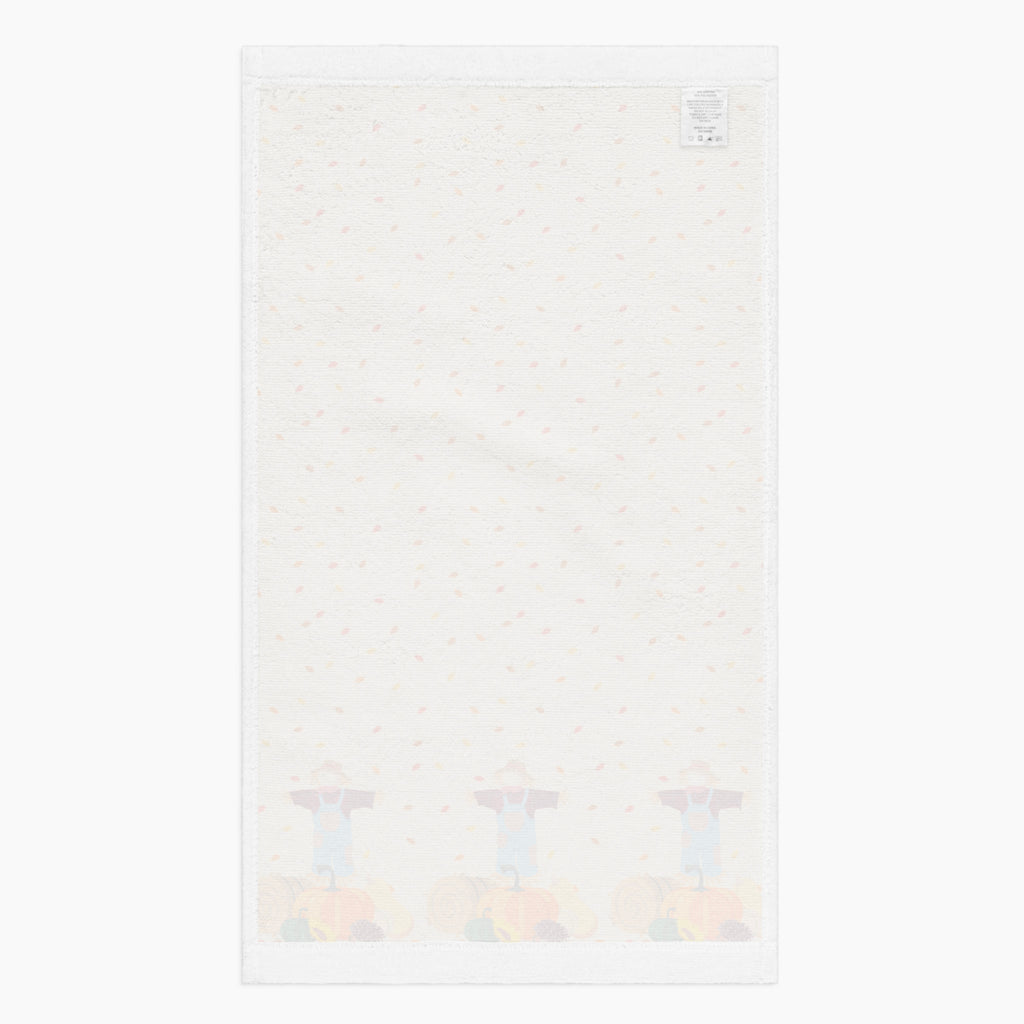A white backside of a rectangular beige kitchen towel, featuring an all-over autumn leaf print and a row of three scarecrows with pumpkins and gourds along the bottom edge.