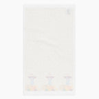 A white backside of a rectangular beige kitchen towel, featuring an all-over autumn leaf print and a row of three scarecrows with pumpkins and gourds along the bottom edge.