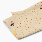 A close-up of a rectangular beige kitchen towel, featuring an all-over autumn leaf print and a row of three scarecrows with pumpkins and gourds along the bottom edge.