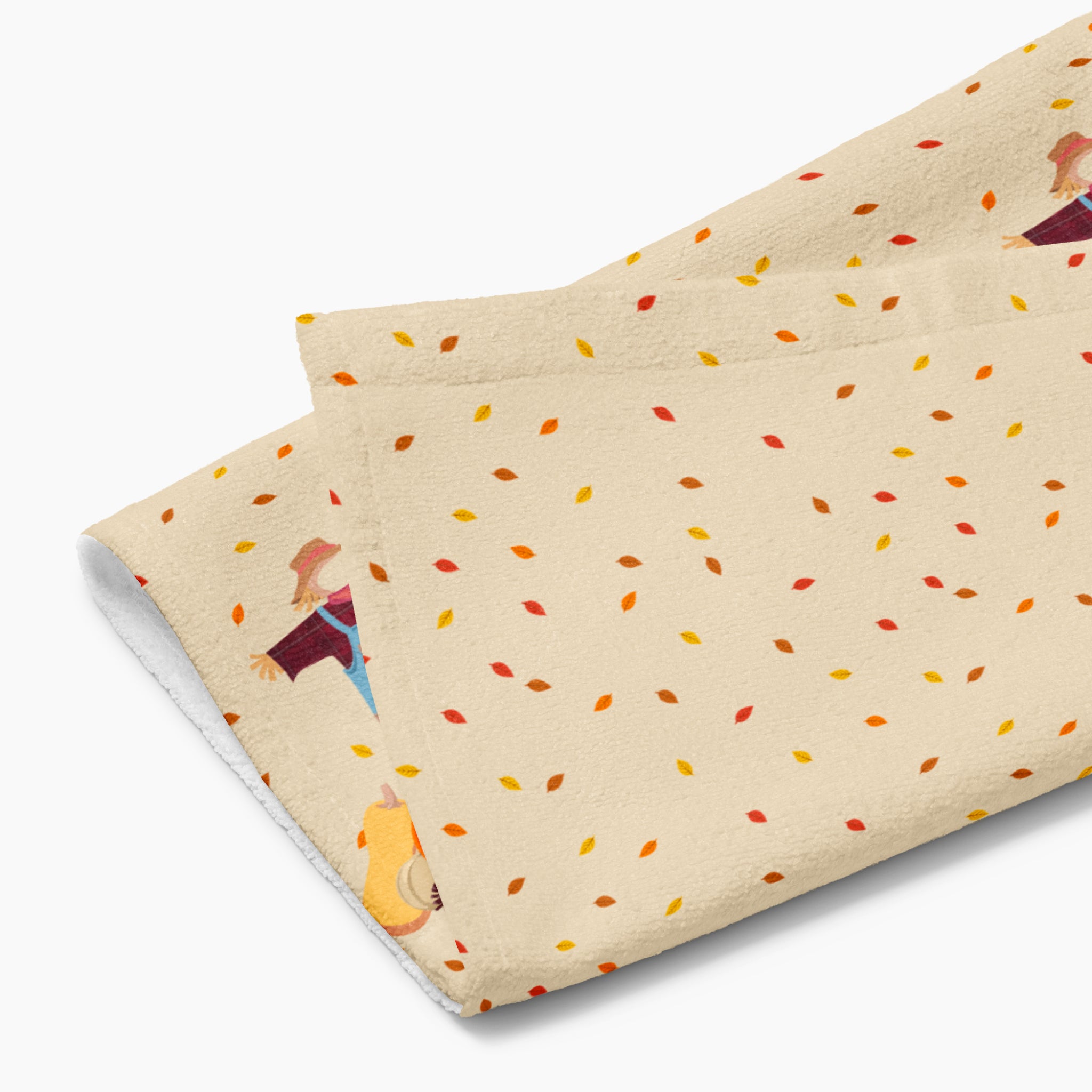 A close-up of a rectangular beige kitchen towel, featuring an all-over autumn leaf print and a row of three scarecrows with pumpkins and gourds along the bottom edge.