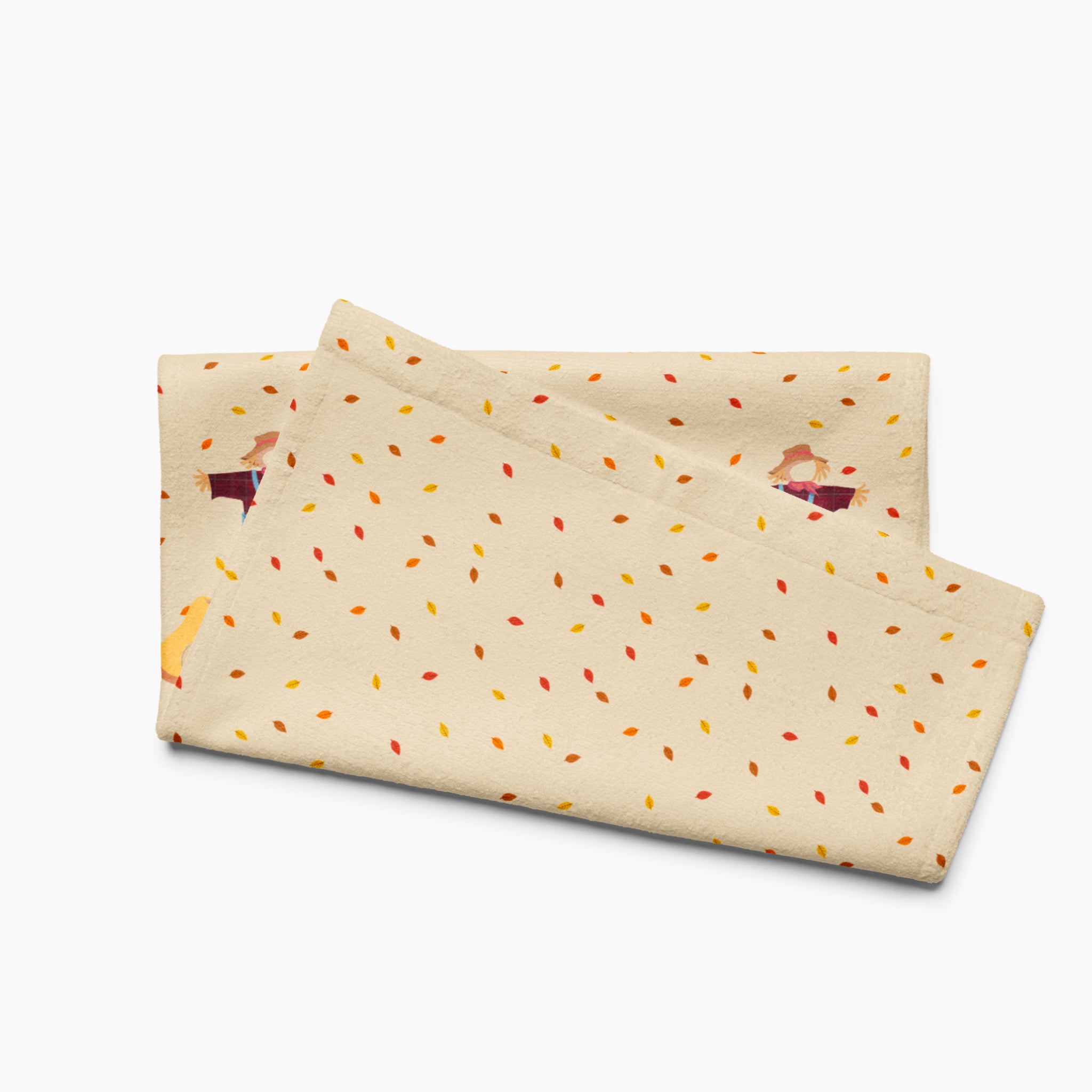 A folded beige kitchen towel, featuring an all-over autumn leaf print and a row of three scarecrows with pumpkins and gourds along the bottom edge.