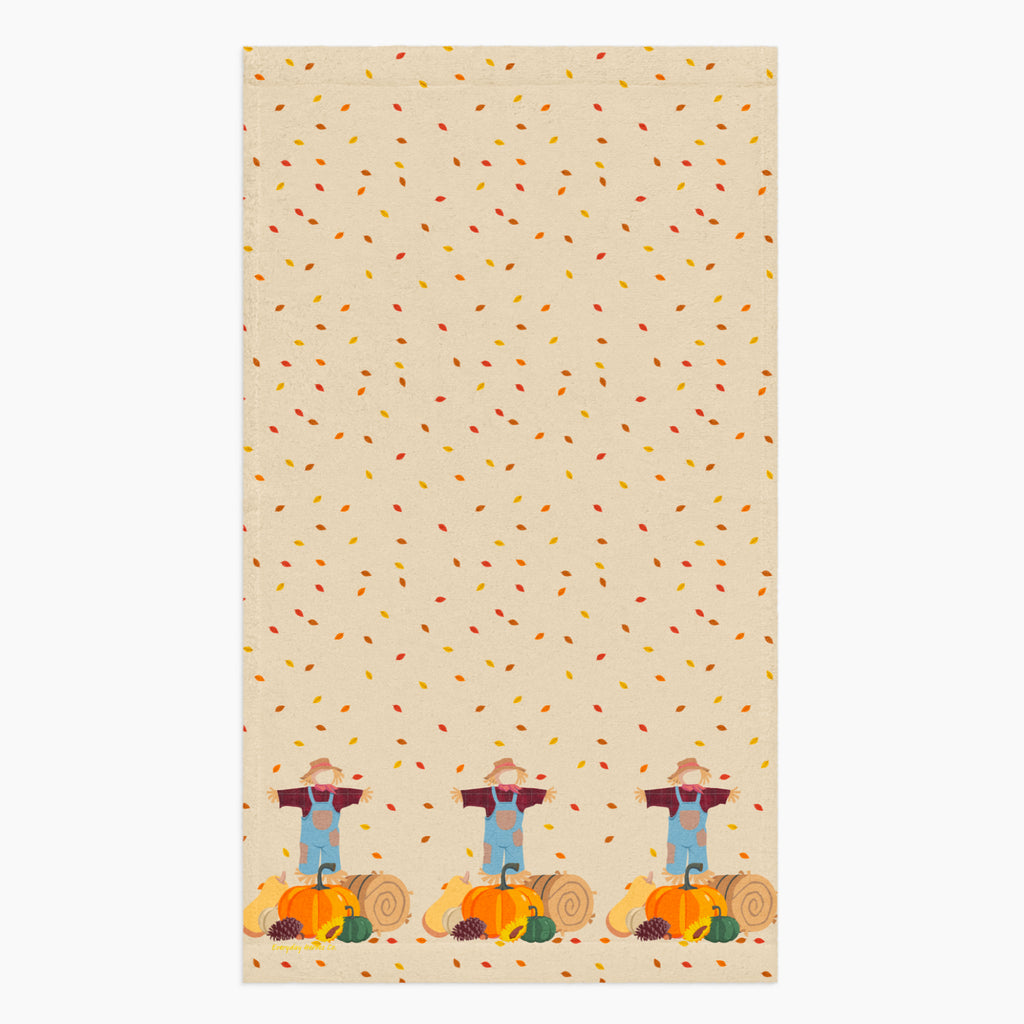 A rectangular beige kitchen towel, featuring an all-over autumn leaf print and a row of three scarecrows with pumpkins and gourds along the bottom edge.