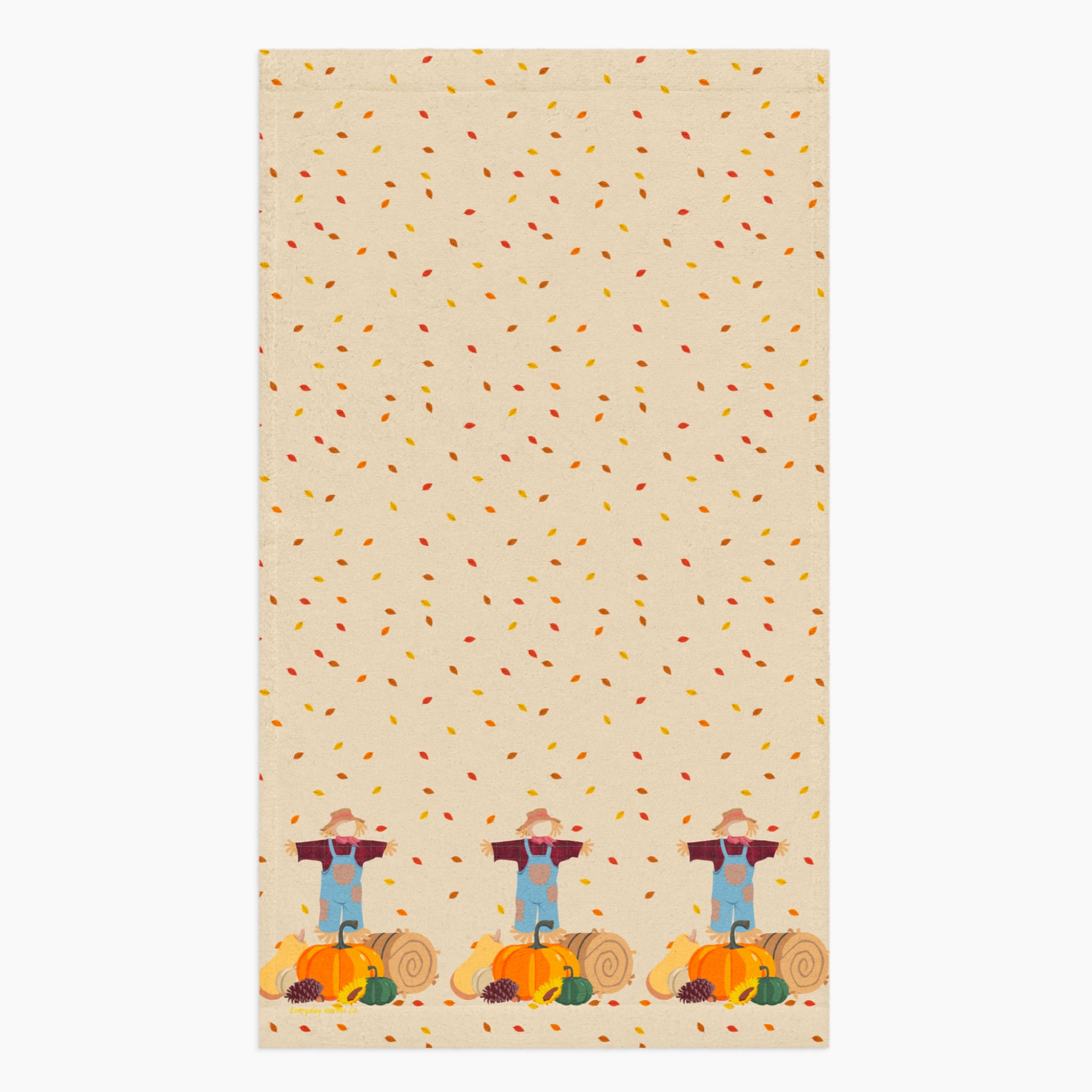 A rectangular beige kitchen towel, featuring an all-over autumn leaf print and a row of three scarecrows with pumpkins and gourds along the bottom edge.