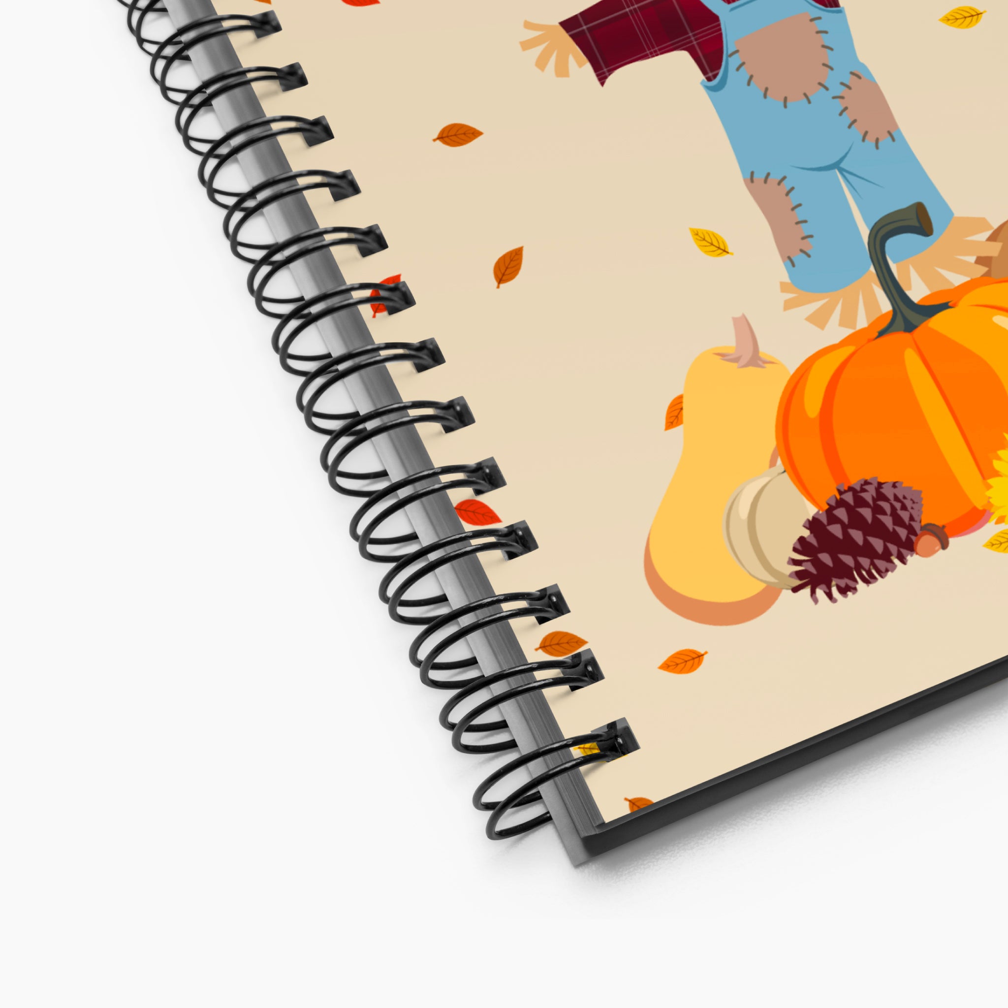 Close-up of a spiral-bound notebook shown at an angle on a white background, featuring a fall harvest illustration with a scarecrow, pumpkins, a gourds, a pinecone, and scattered autumn leaves.