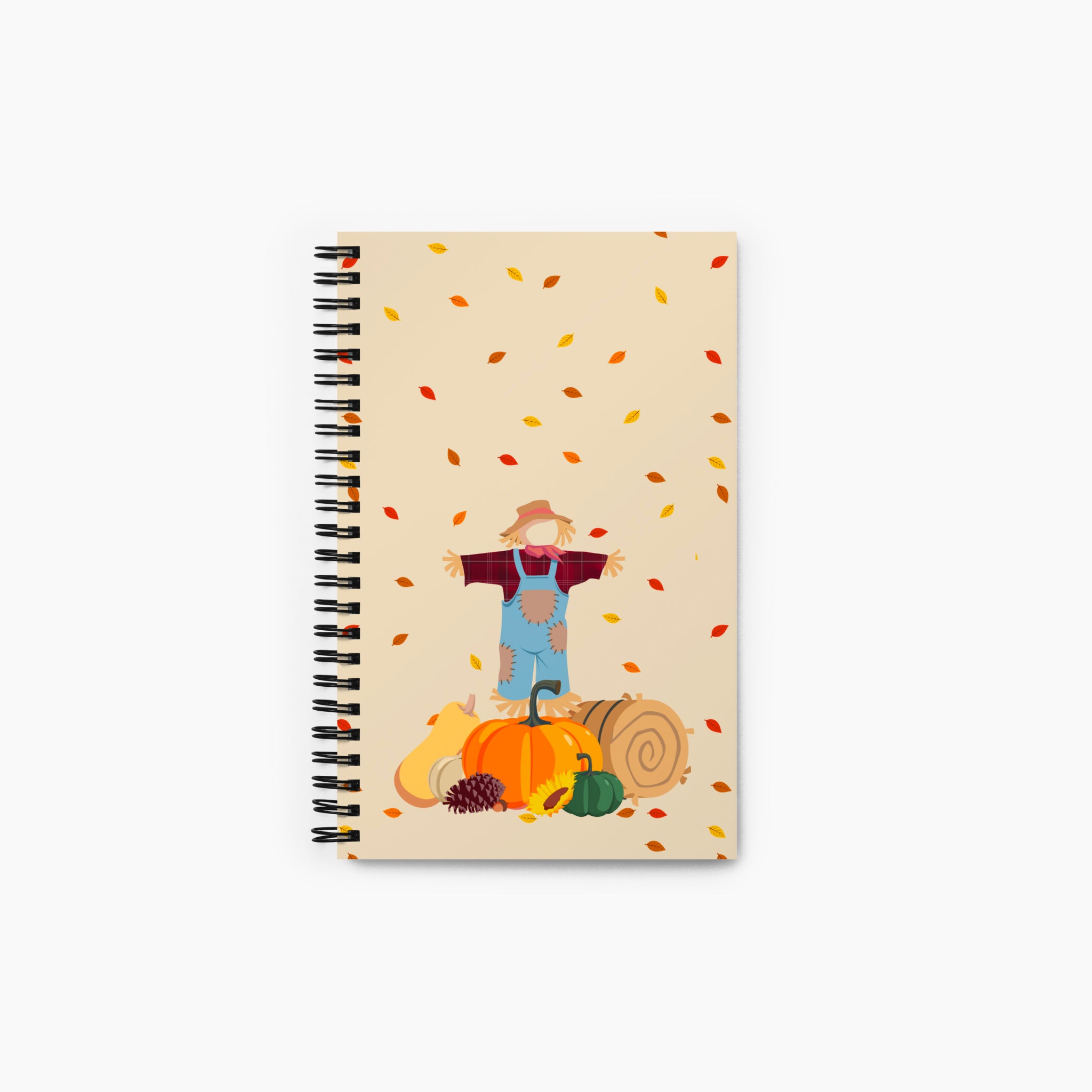 A spiral-bound notebook with a beige cover featuring a cartoon scarecrow, pumpkins, gourds, and falling autumn leaves, shown on a light gray background.